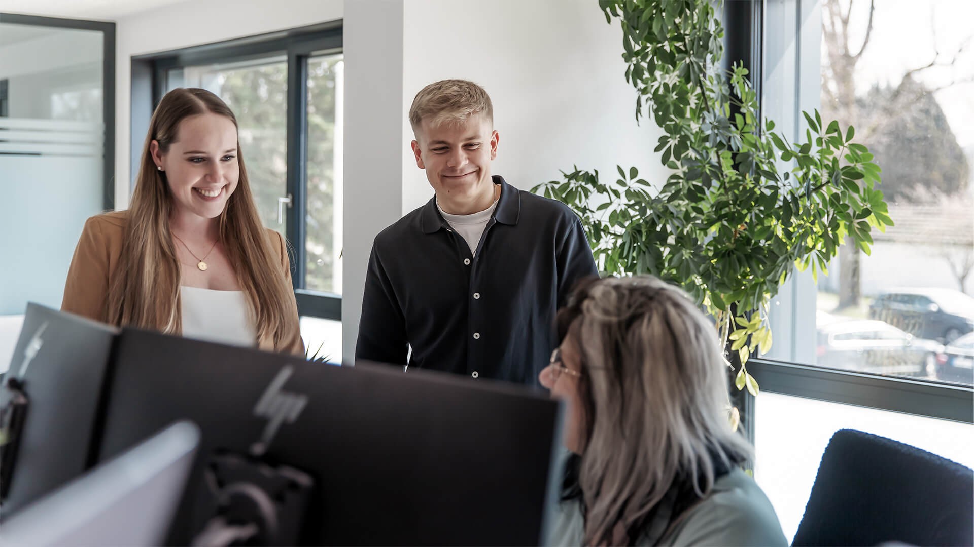 Foto: Drei Personen in einem modernen Büro im Gespräch. Eine Frau mit langen braunen Haaren und braunem Blazer sowie ein junger Mann im dunklen Hemd stehen nebeneinander und lächeln. Ihnen gegenüber sitzt eine Frau mit grauen Haaren an einem Schreibtisch vor zwei Monitoren. Im Hintergrund sind große Fenster und eine grüne Pflanze zu sehen.