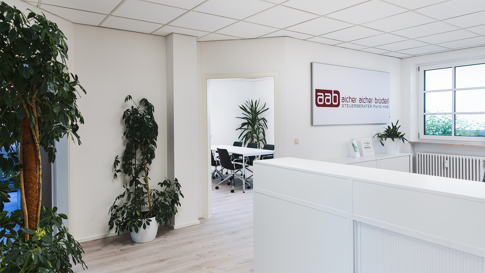 Foto: Heller Empfangsbereich des Büros von Aicher Aicher Brüderl Steuerberater PartG mbB   mit weißem Tresen. An der Wand hängt ein Schild mit der Aufschrift "Aab Aicher Aicher Brüderl Steuerberater PartG mbB". Neben dem Fenster und auf dem Tresen stehen grüne Pflanzen. Links im Bild sind weitere große Pflanzen zu sehen. Durch eine offene Tür blickt man in einen Besprechungsraum mit Tisch und schwarzen Stühlen.