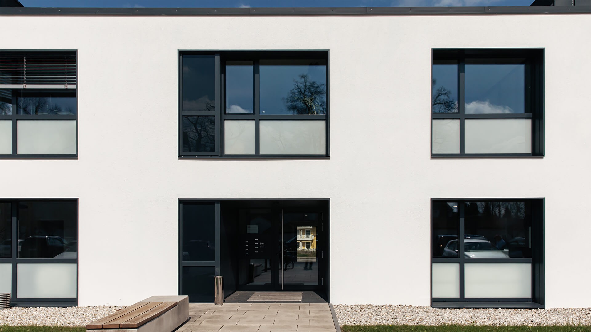 Foto: Moderne Bürogebäude-Fassade von	Aicher Aicher Brüderl Steuerberater PartG mbB  in weiß mit großen dunklen Fenstern. In der Mitte befindet sich der Eingang mit Glas­tür. Vor dem Gebäude stehen eine lange Holzbank und Kiesflächen, im Vordergrund ist eine Rasenfläche zu sehen.